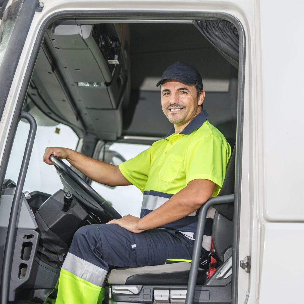 Smiling male driver sitting in truck and looking at camera Positive male truck driver in lemon and blue uniform smiling and looking at camera while sitting in truck with open door on bright day