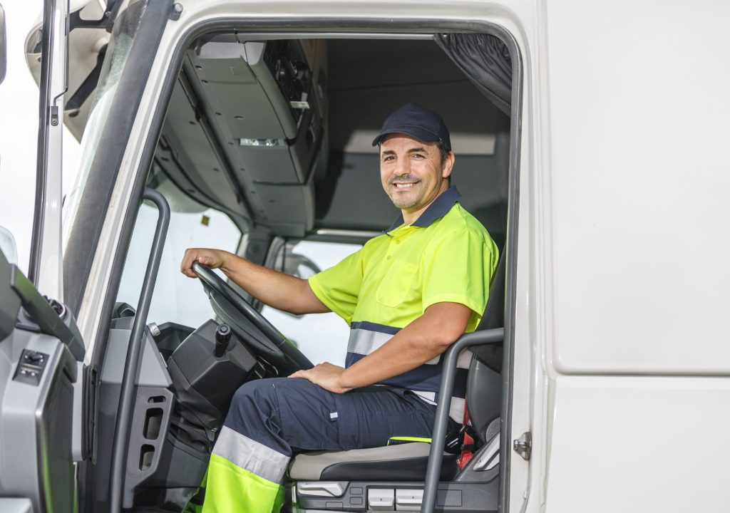 Smiling male driver sitting in truck and looking at camera Positive male truck driver in lemon and blue uniform smiling and looking at camera while sitting in truck with open door on bright day